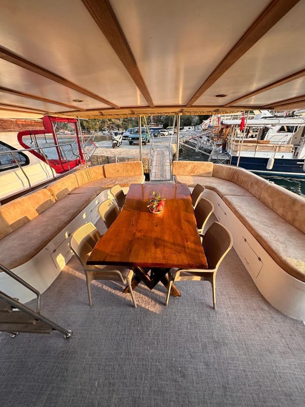 The aft deck at golden hour: a long wooden table, cushions all round, the gangway down to shore